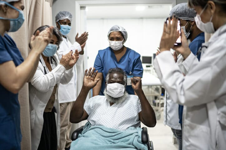 A diverse group of healthcare workers celebrate a senior man leaving the hospital in a wheelchair