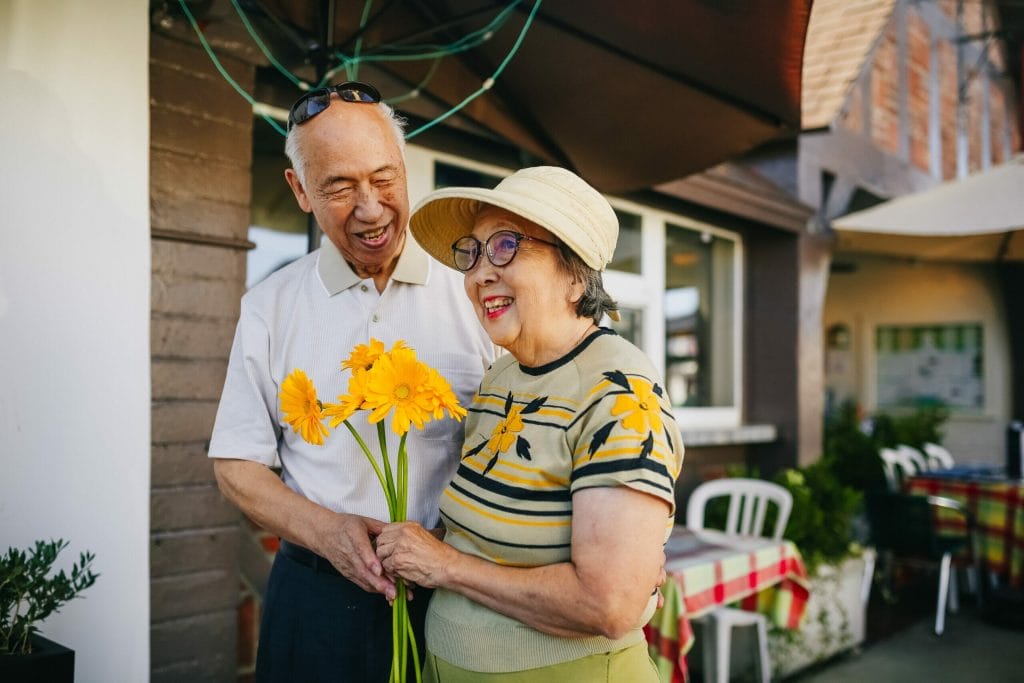 Silver tsunami. An older couple stand outside together, smiling. The woman holds yellow flowers.